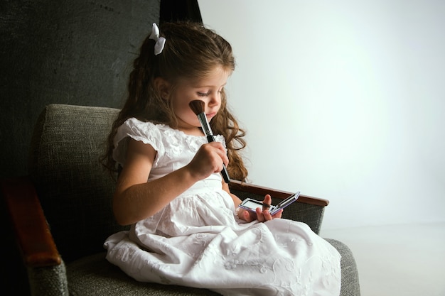 Interação das crianças com o mundo adulto. Linda garota tentando fazer brilhante compensar por ser mais velha. Modelo feminino experimentando os cosméticos da mãe em casa. Infância, estilo, moda, conceito de sonho.