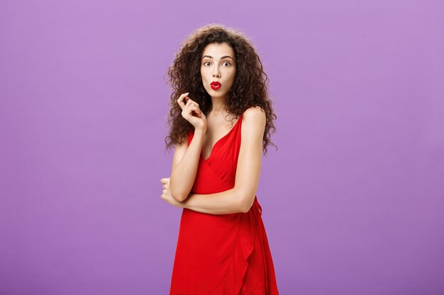 Intrigada chaming senhora elegante com penteado encaracolado em vestido vermelho e batom dobrando os lábios curiosamente ...