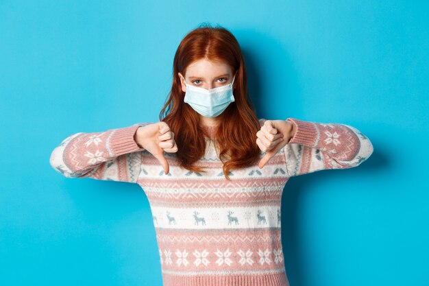 Inverno, covid-19 e conceito de pandemia. Garota ruiva chateada e com raiva na máscara facial mostrando desaprovação, polegares para baixo em antipatia, em pé sobre um fundo azul.