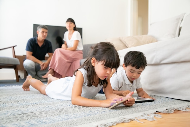 Irmão e irmãzinha fofos usando aplicativos de aprendizagem em dispositivos, deitados no chão enquanto os pais sentam juntos