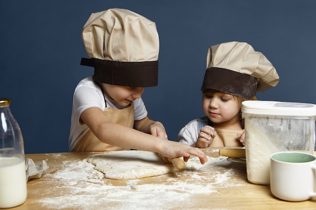 Irmãos adoráveis menino e menina fazendo biscoitos juntos, de pé na mesa da cozinha com uma garrafa de leite, farinha, achatando a massa usando o rolo. Família, infância, padaria caseira, alegria e felicidade