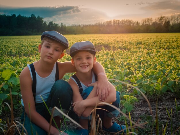 Irmãos com chapéus e suspensórios em um campo coberto de vegetação durante o pôr do sol