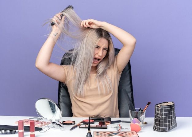 Irritada com os olhos fechados, uma jovem linda se senta à mesa com ferramentas de maquiagem, penteando o cabelo isolado na parede azul