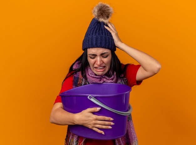 Irritada e dolorida, jovem caucasiana doente usando chapéu de inverno e lenço segurando um balde de plástico, olhando para dentro, tendo náuseas isoladas em um fundo laranja com espaço de cópia
