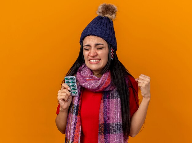 Irritada, jovem caucasiana doente com chapéu de inverno e lenço segurando o pacote de cápsulas com outros pacotes sob o chapéu cerrando o punho com os olhos fechados, isolado na parede laranja com espaço de cópia