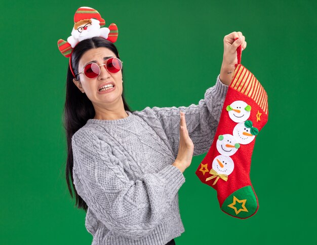 Irritada, jovem caucasiana, usando uma faixa de papai noel com óculos segurando uma meia de natal, olhando para a câmera, fazendo gesto de recusa isolado no fundo verde