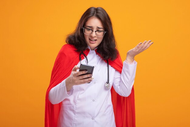 Irritada, jovem super-heroína caucasiana vestindo uniforme de médico e estetoscópio com óculos segurando e olhando para o telefone celular, mostrando a mão vazia isolada na parede