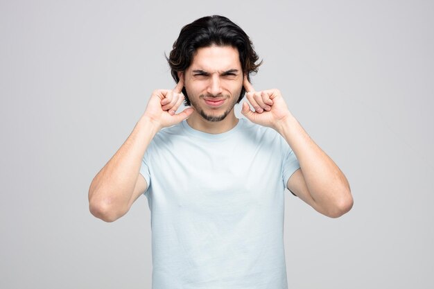 irritado jovem bonito colocando os dedos nos ouvidos olhando para câmera isolada no fundo branco