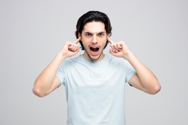 Irritado jovem bonito olhando para a câmera colocando os dedos nos ouvidos gritando isolado no fundo branco