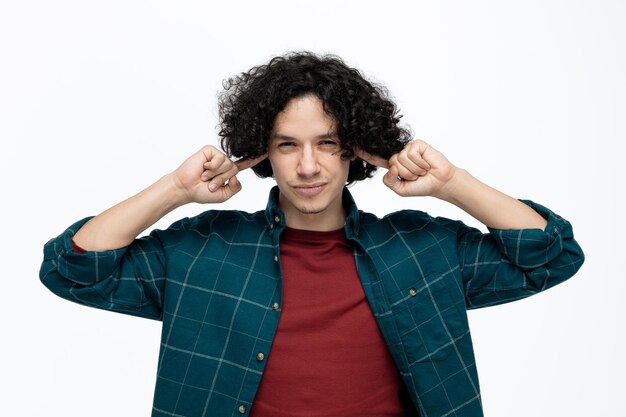 Irritado jovem bonito olhando para a câmera fechando os ouvidos com os dedos isolados no fundo branco