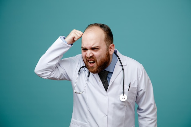 Irritado jovem médico masculino vestindo jaleco e estetoscópio em volta do pescoço, olhando para o lado mostrando você é um gesto idiota isolado em fundo azul