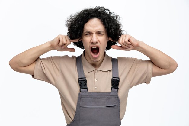 Foto grátis irritado jovem trabalhador da construção civil vestindo uniforme olhando para a câmera colocando os dedos nos ouvidos gritando isolado no fundo branco