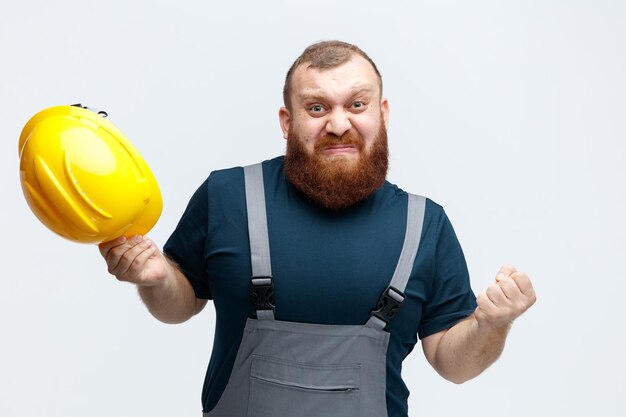 Irritado jovem trabalhador da construção civil vestindo uniforme segurando capacete de segurança mantendo o punho no ar olhando para câmera isolada no fundo branco