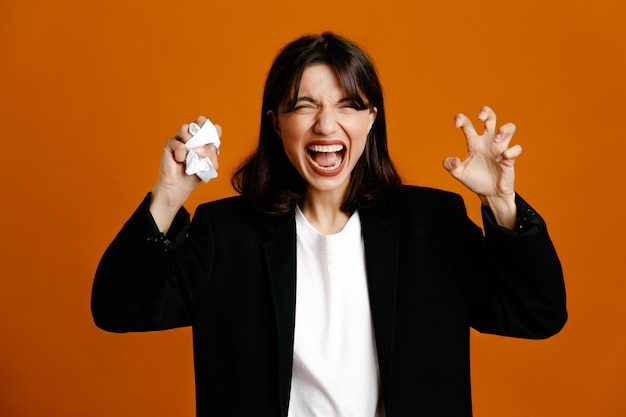 Irritado mostrando gesto de estilo tigre rasgando papel jovem linda fêmea vestindo jaqueta preta isolada em fundo laranja