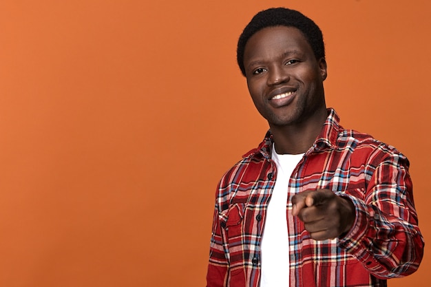 Isolado bonito alegre jovem negro de pele escura vestindo camisa xadrez elegante, sorrindo amplamente e apontando o dedo da frente como se estivesse escolhendo você. foco seletivo