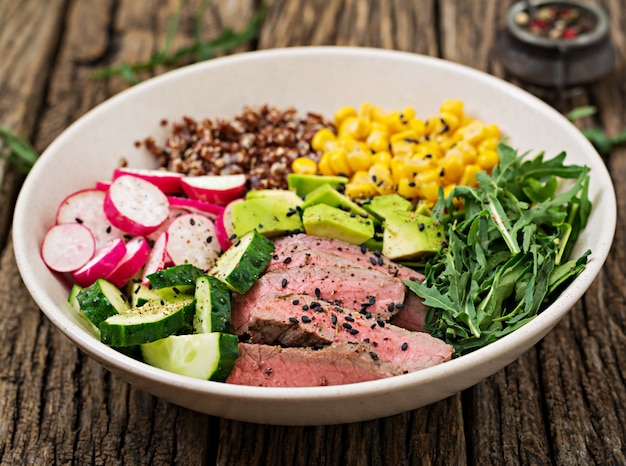 Jantar saudável. almoço de tigela de buda com bife grelhado e quinoa, milho, abacate, pepino e rúcula na mesa de madeira. salada de carne.