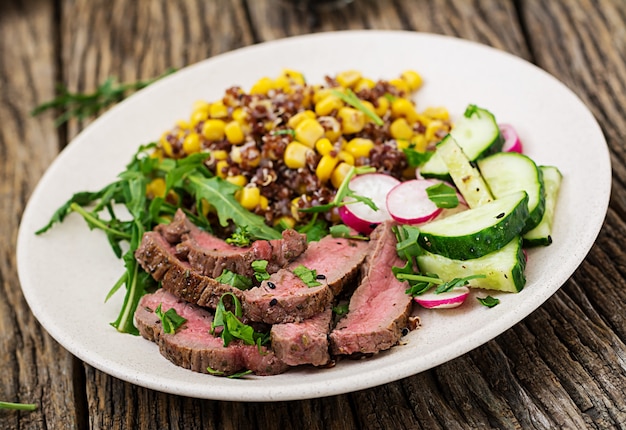 Jantar saudável. Tigela de almoço com bife grelhado e quinoa, milho, pepino, rabanete e rúcula na mesa de madeira. Salada de carne.