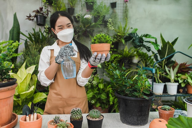 Jardineira asiática usando máscara e avental segurando um spray para regar uma planta de casa verde
