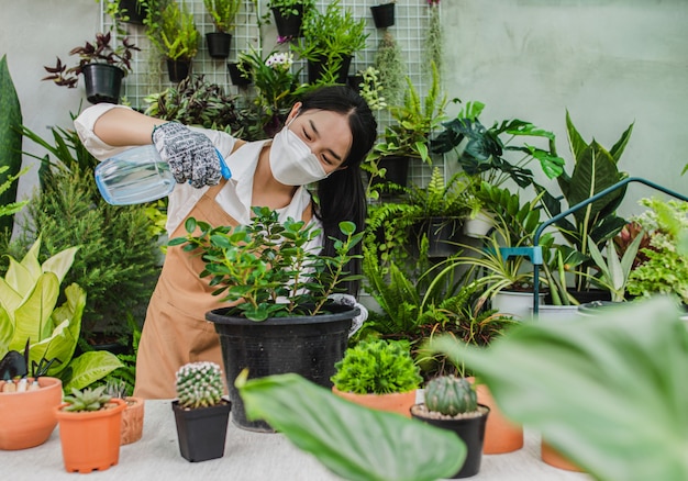 Jardineira asiática usando máscara e avental segurando um spray para regar uma planta de casa verde