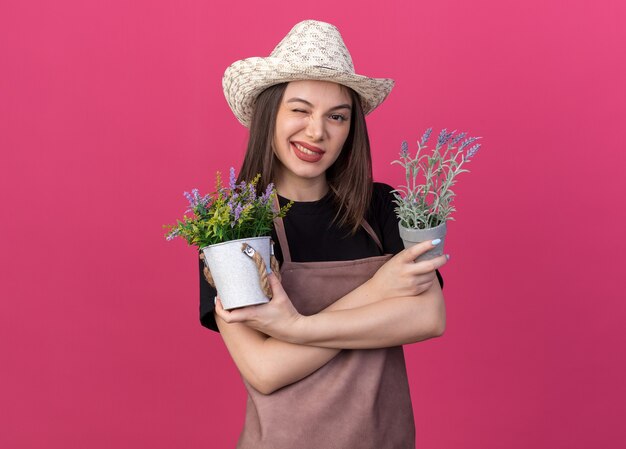 Jardineira confiante, bonita, caucasiana, usando chapéu de jardinagem, pisca os olhos e fica de pé com os braços cruzados segurando vasos de flores rosa