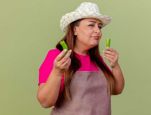 Jardineira de meia idade com avental e chapéu mostrando metades de pimenta verde olhando para a câmera com uma expressão cética de pé sobre um fundo claro