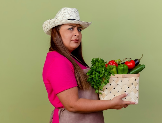 Jardineira de meia-idade com avental e chapéu segurando uma caixa cheia de legumes
