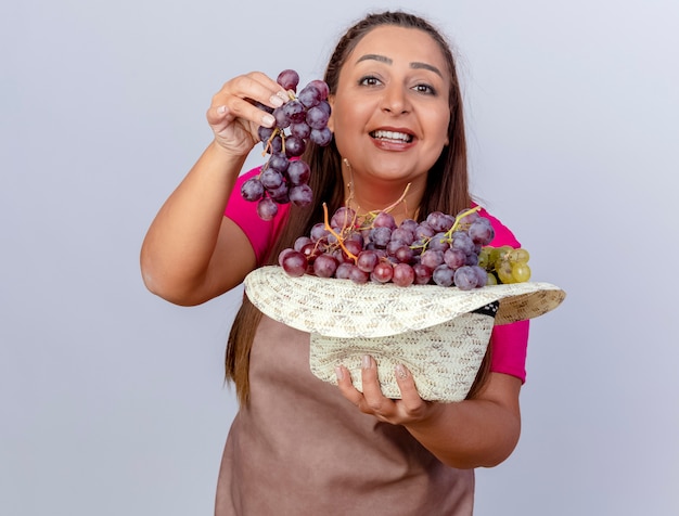 Jardineira de meia-idade com avental segurando um chapéu cheio de uvas com um sorriso no rosto