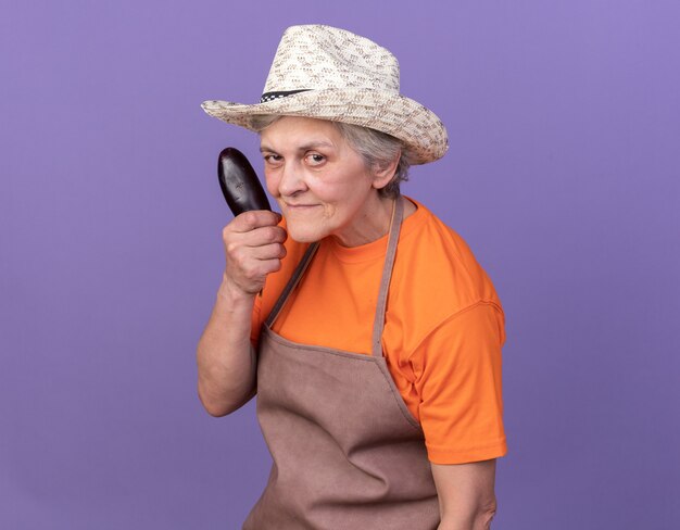 Jardineira idosa confiante usando chapéu de jardinagem segurando berinjela
