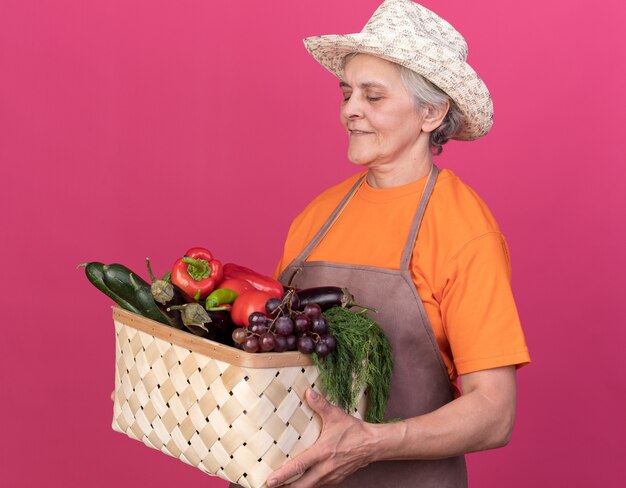 Jardineira idosa satisfeita com chapéu de jardinagem, segurando e olhando para a cesta de vegetais