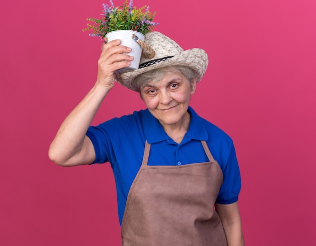 Jardineira idosa satisfeita com um chapéu de jardinagem segurando um vaso de flores sobre a cabeça