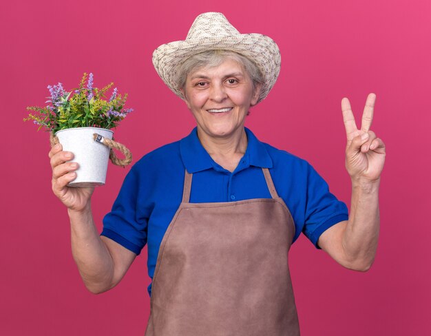 Jardineira idosa sorridente usando chapéu de jardinagem segurando um vaso de flores e gesticulando a placa da vitória rosa