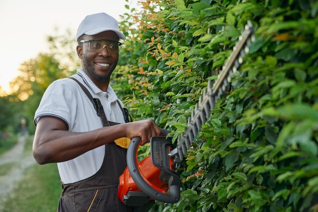 Jardineiro afro sorridente usando aparador de cerca viva para cortar arbustos