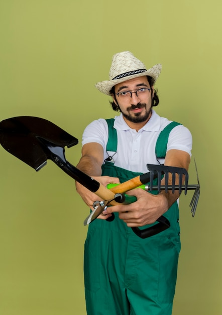 Jardineiro confiante usando óculos óticos e chapéu de jardinagem segurando ferramentas de jardim