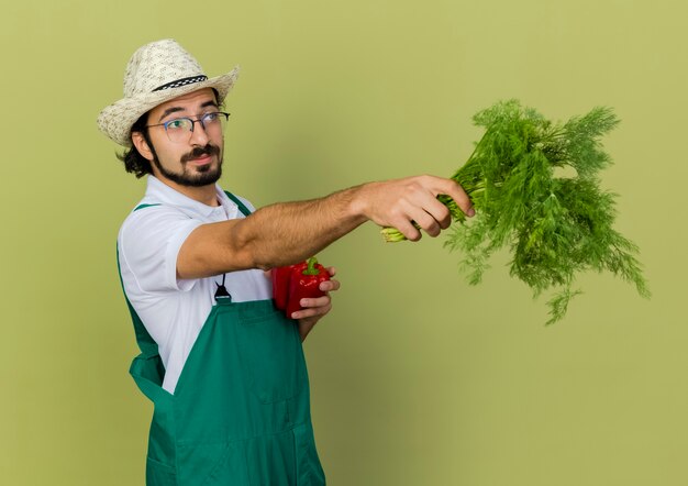 Jardineiro confiante usando óculos óticos e chapéu de jardinagem segurando pimentas vermelhas