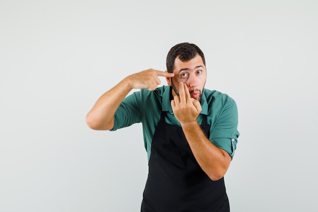 Jardineiro masculino apontando para a pálpebra puxada pelo dedo na camiseta, vista frontal do avental.