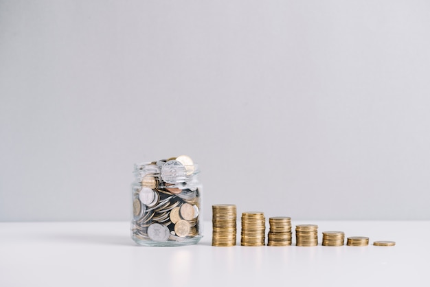 Jarra de vidro cheia de dinheiro na frente de diminuir moedas empilhadas contra um fundo branco