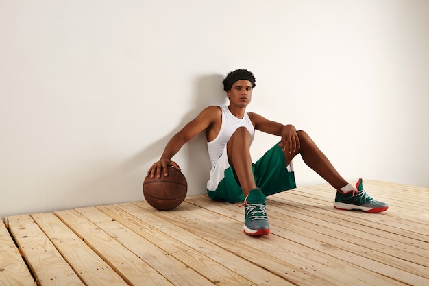 Jogador de basquete preto cansado e pensativo em outift de basquete verde e branco, sentado no chão de madeira clara, descansando a mão em uma bola de basquete marrom grunge