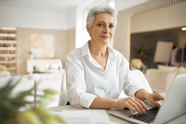 jornalista mulher madura com cabelo grisalho curto digitando no laptop, sentada no local de trabalho com as mãos no teclado, inspirando a expressão facial