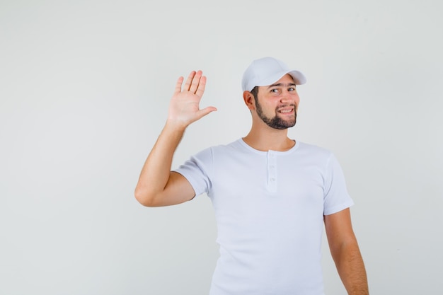 Jovem acenando com a mão para saudação em t-shirt branca, boné e olhando fresco, vista frontal.