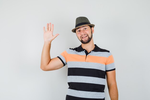 Jovem acenando com a mão para saudação em t-shirt, chapéu e parece alegre.