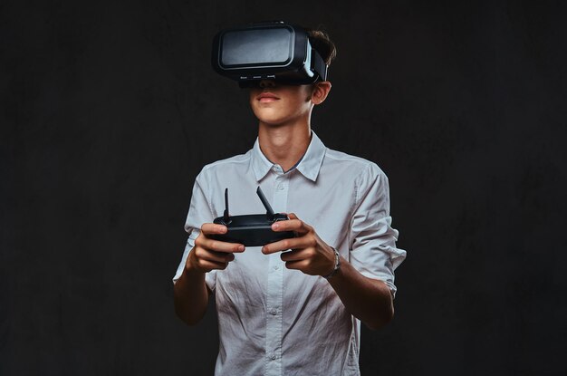 Jovem adolescente vestido com uma camisa branca usa óculos de realidade virtual e controla o quadcopter usando o controle remoto.