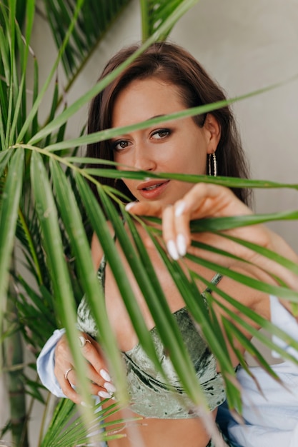 Foto grátis jovem adorável com grandes olhos lindos cobrindo por folha de palmeira e posando para a câmera usando um top e uma camisa da moda. lugar para texto