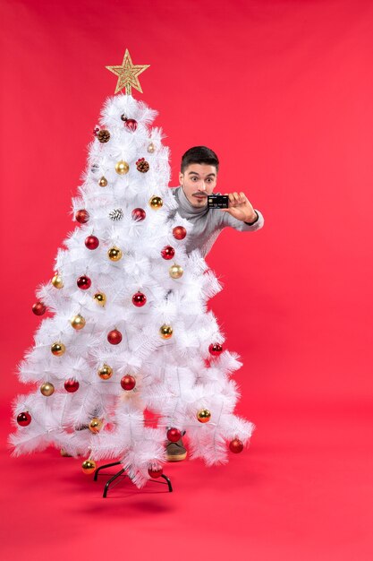 Jovem adulto chocado com uma blusa cinza em pé atrás da árvore de Natal decorada e olhando para o telefone vermelho