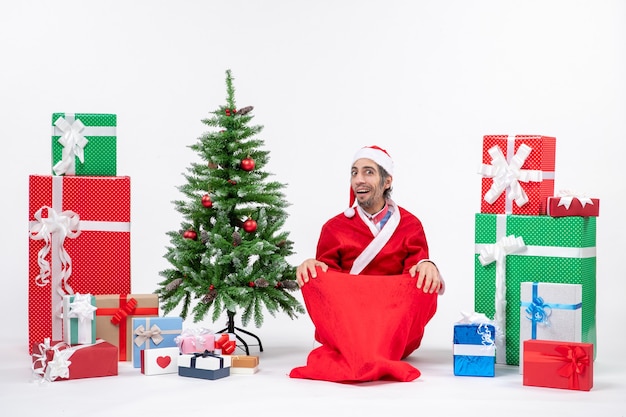 Jovem adulto chocado vestido de papai noel com presentes e uma árvore de natal decorada sentada no chão sobre um fundo branco