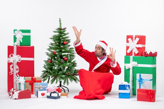Jovem adulto engraçado vestido de papai noel com presentes e uma árvore de natal decorada, sentado no chão apontando para cima, fazendo gesto de óculos no fundo branco
