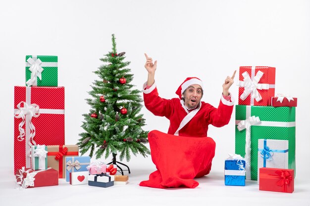 Jovem adulto surpreso vestido de Papai Noel com presentes e uma árvore de Natal decorada sentado no chão apontando algo em ambos os lados no fundo branco