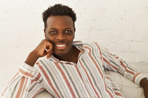 Jovem africano alegre e atraente em uma camisa listrada, sentado confortavelmente no sofá da sala de estar, colocando a mão no queixo e olhando para a câmera com um sorriso largo e radiante