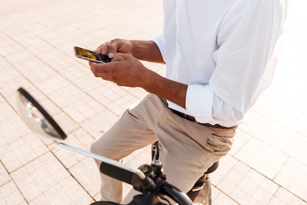 jovem Africano sentado na moto moderna ao ar livre e usando smartphone