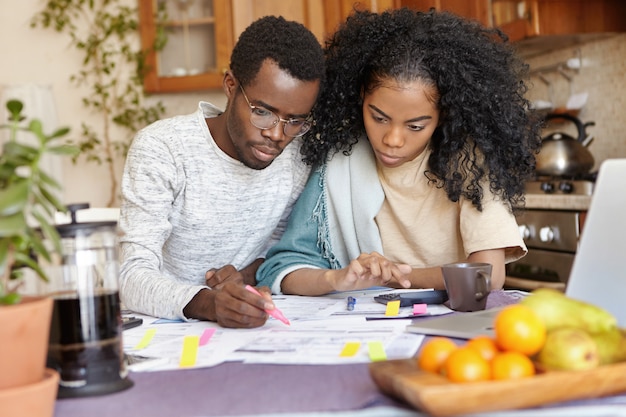 Jovem africano sério de óculos, segurando uma caneta de feltro, calculando as despesas domésticas enquanto administrava o orçamento familiar junto com sua linda esposa, planejando uma grande compra e tentando economizar dinheiro