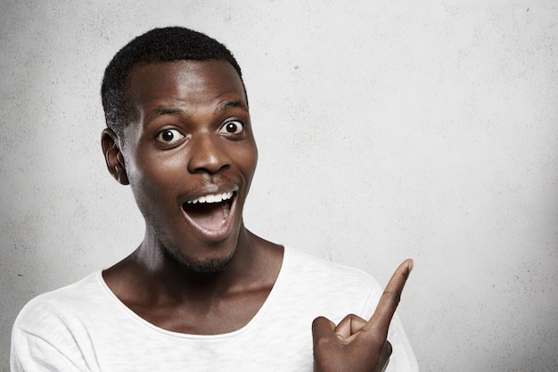 Foto grátis jovem africano vestido com uma camiseta branca, abrindo a boca amplamente em surpresa, parecendo chocado com os preços de venda altos, mostrando algo na parede em branco com o dedo.